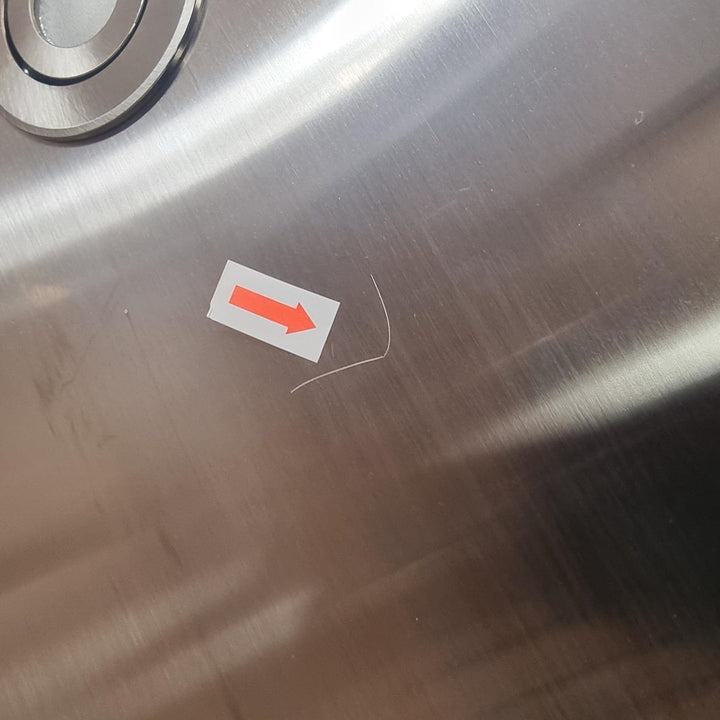 A close-up of a brushed stainless steel surface with a small white sticker featuring a red arrow pointing right, placed near a scratch on the metal.