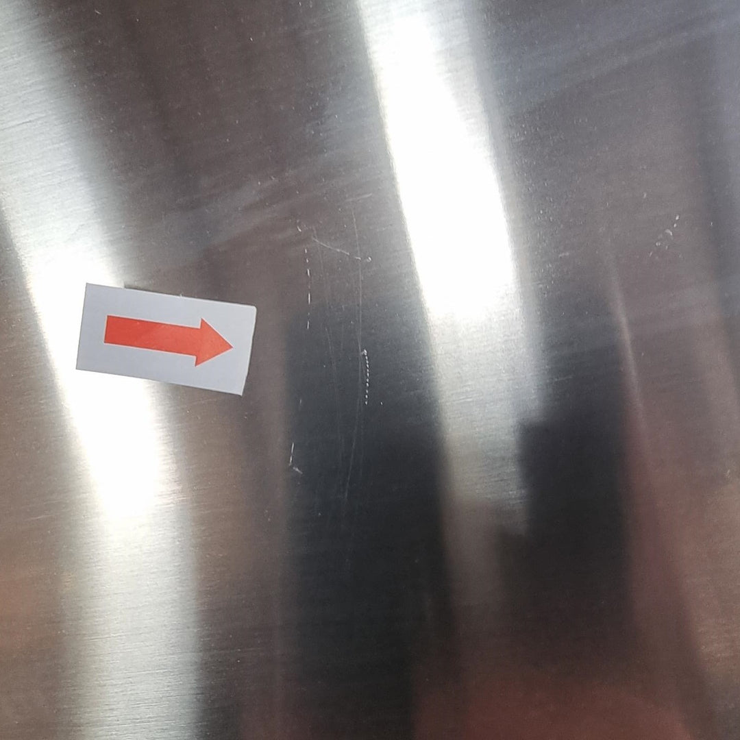 A close-up of a brushed stainless steel surface with a small white sticker featuring a red right-pointing arrow, plus visible scratches