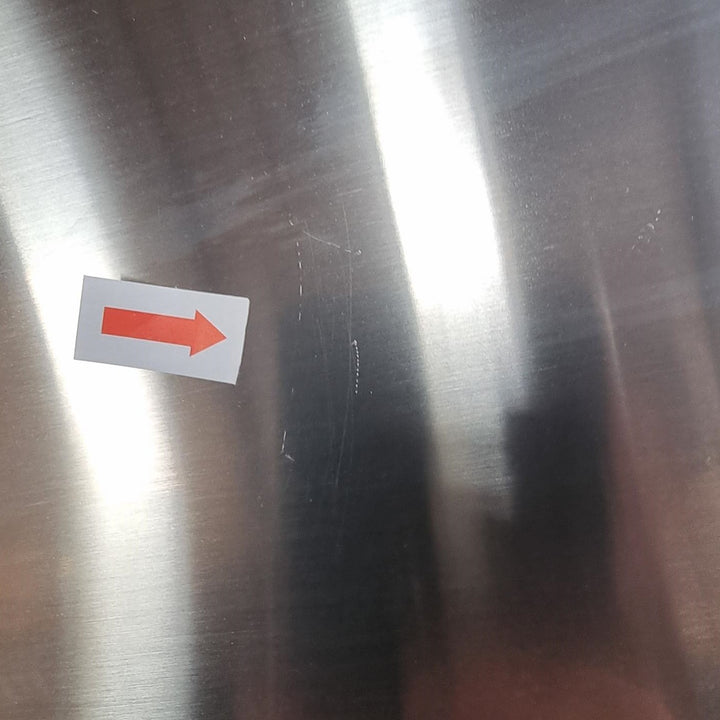 A close-up of a brushed stainless steel surface with a small white sticker featuring a red right-pointing arrow, plus visible scratches