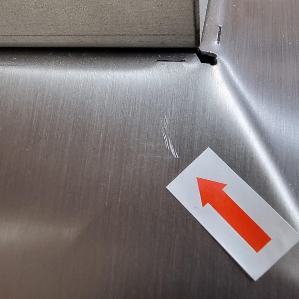 A close-up of a brushed stainless steel surface with a small white rectangular sticker featuring a red arrow pointing upward, near the corner of another metal panel.
