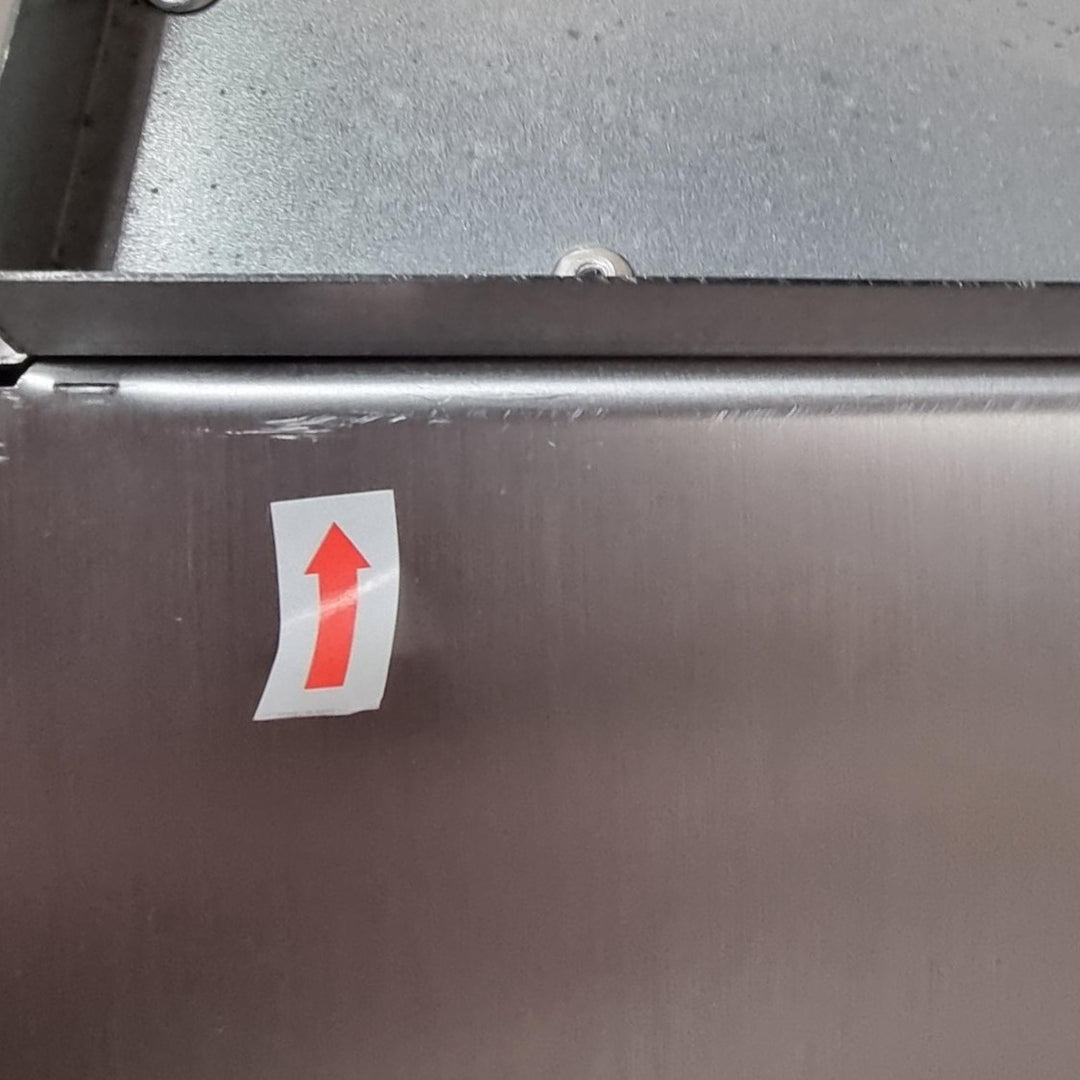 Close-up of a brushed metal surface with minor scratches with a small white sticker featuring a red upward-pointing arrow near the centre-left.
