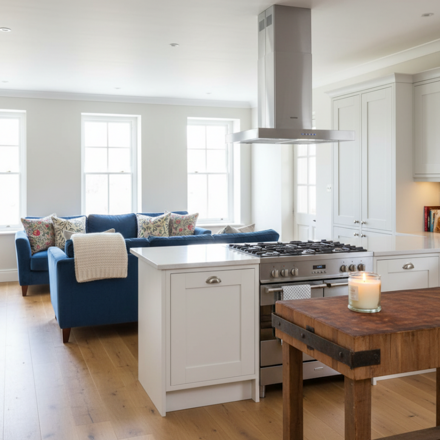 90cm Flat island cooker hood in stainless steel above kitchen island and stove, adjacent to a living room with blue sofa and floral pillows.