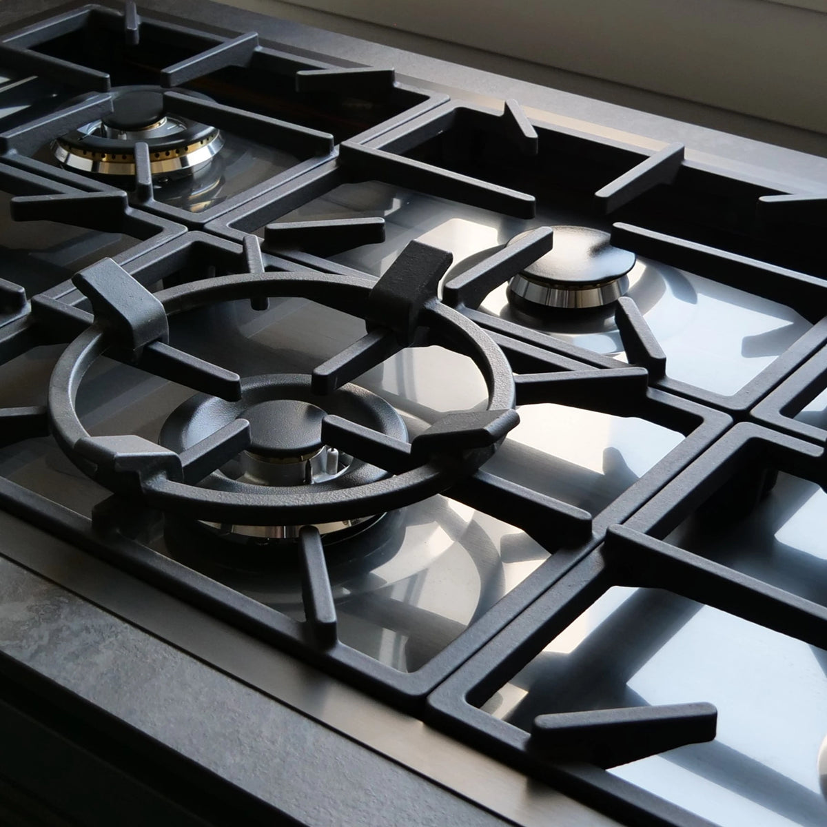Close-up of a gun metal grey gas hob with multiple burner grates and metallic burner centres on a dark cooktop surface.