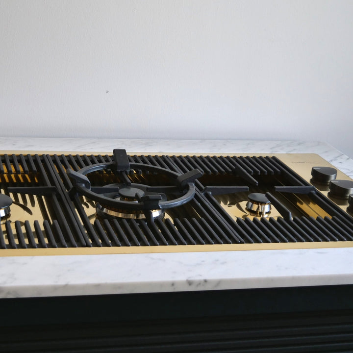 A gold 87cm 3 burner gas hob with black grates and knobs set into a white marble countertop with a brushed gold metal faceplate.
